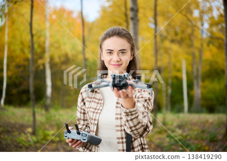 A cheerful young woman stands in a vibrant autumn forest, holding a drone and its remote control, ready to take flight amidst the golden leaves. A cheerful young woman stands in a vibrant autumn forest, holding a drone and its remote control, ready to take flight amidst the golden leaves. 138419290
