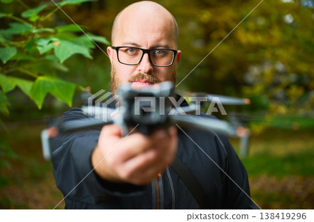 A man stands confidently in a lush green park, holding a drone in front of him. The vibrant fall foliage surrounds him, showcasing the beauty of nature while he prepares to fly. A man stands confidently in a lush green park, holding a drone in front of him. The vibrant fall foliage surrounds him, showcasing the beauty of nature while he prepares to fly. 138419296