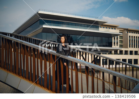 Woman stands on bridge near modern building in the city during sunny day with clear sky Woman stands on bridge near modern building in the city during sunny day with clear sky 138419506