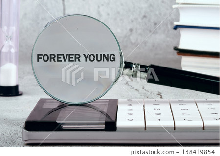 Magnifier lens reveals FOREVER YOUNG on a calculator. A slender hourglass marks times gentle flow, poised on a textured desk next to stacked books Magnifier lens reveals FOREVER YOUNG on a calculator. A slender hourglass marks times gentle flow, poised on a textured desk next to stacked books 138419854