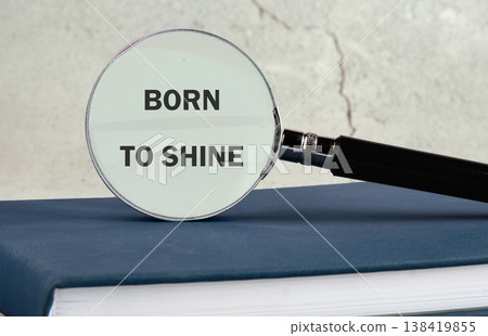 A magnifying glass rests upon a blue book, highlighting the empowering phrase Born To Shine against a textured background A magnifying glass rests upon a blue book, highlighting the empowering phrase Born To Shine against a textured background 138419855