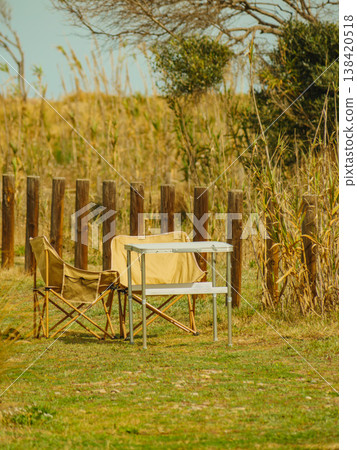 Two chairs and a table are set up in an open field beside wooden posts with tall grass and bushes in the background Two chairs and a table are set up in an open field beside wooden posts with tall grass and bushes in the background 138420518