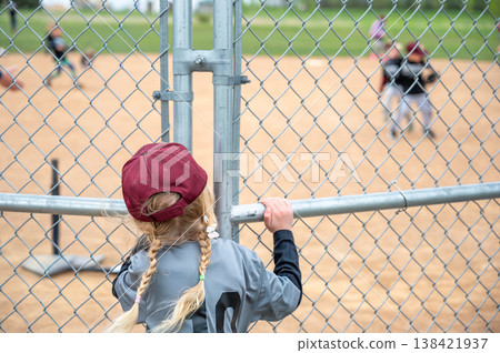 Girl watches baseball game through fence at local park during spring afternoon Girl watches baseball game through fence at local park during spring afternoon 138421937