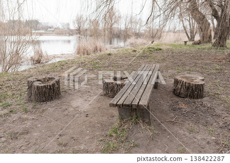 Rustic Wooden Bench and Tree Stump Seats Beside Tranquil Lake With Bare Trees In Early Spring Landscape 138422287