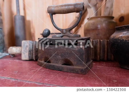 Vintage Rusty Cast Iron Sad Iron With Wooden Handle Displayed On Red Table Among Antique Kitchen Utensils And Ceramic Jars In Warm Wood Background 138422308