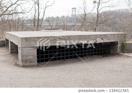 Weathered Concrete Shelter with Black Metal Grate Ventilation System Against Bare Trees and Distant Suspension Bridge in Gray Winter Landscape Weathered Concrete Shelter with Black Metal Grate Ventilation System Against Bare Trees and Distant Suspension Bridge in Gray Winter Landscape 138422365