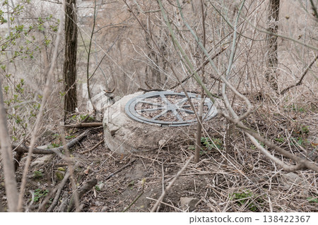 Abandoned Metal Manhole Cover Resting on Concrete Block Amidst Overgrown Winter Forest With Bare Trees and Dry Brush Under Natural Light Abandoned Metal Manhole Cover Resting on Concrete Block Amidst Overgrown Winter Forest With Bare Trees and Dry Brush Under Natural Light 138422367