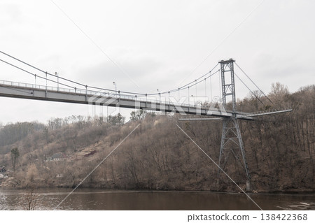 Gray Suspension Bridge Spanning River Amidst Bare Forest Hills Under Overcast Sky With Visible Steel Tower Structure And Calm Water Surface Below Gray Suspension Bridge Spanning River Amidst Bare Forest Hills Under Overcast Sky With Visible Steel Tower Structure And Calm Water Surface Below 138422368