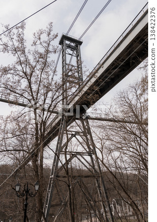 Industrial Steel Suspension Bridge Tower Amidst Bare Trees Under Overcast Sky With Vintage Street Lamp In Foreground And Gray Cloudy Background 138422376