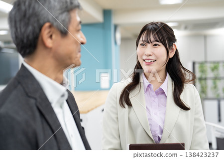 Two office workers, holding documents and laptops, are chatting in a cheerful manner. Two office workers, holding documents and laptops, are chatting in a cheerful manner. 138423237
