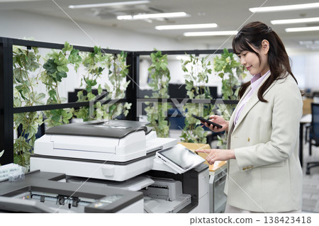 一名女員工在辦公室用智慧型手機操作多功能印表機。 一名女員工在辦公室用智慧型手機操作多功能印表機。 138423418