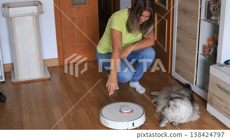 Woman in a green shirt kneels beside a robotic vacuum cleaner on wooden flooring, while a fluffy cat sits nearby Woman in a green shirt kneels beside a robotic vacuum cleaner on wooden flooring, while a fluffy cat sits nearby 138424797
