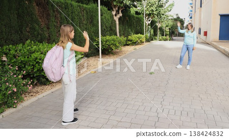 A mother and her school-age daughter wave farewell outside the school entrance. Back to school vibes A mother and her school-age daughter wave farewell outside the school entrance. Back to school vibes 138424832