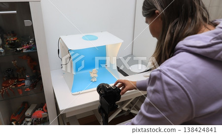 Person takes a picture of a small figure in a light box with a blue background inside a room filled with toys and small items in the background Person takes a picture of a small figure in a light box with a blue background inside a room filled with toys and small items in the background 138424841