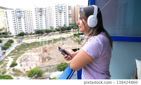 Woman with headphones listens to music on smartphone while standing on balcony Woman with headphones listens to music on smartphone while standing on balcony 138424860