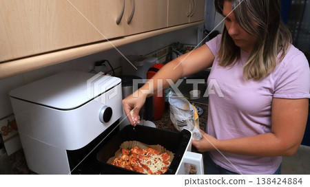 Woman in kitchen using air fryer to prepare meal, adding seasoning to dish  138424884