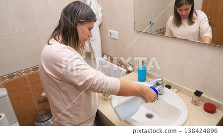 Woman cleaning bathroom sink with spray bottle in a tiled bathroom, mirror reflecting her actions Woman cleaning bathroom sink with spray bottle in a tiled bathroom, mirror reflecting her actions 138424896