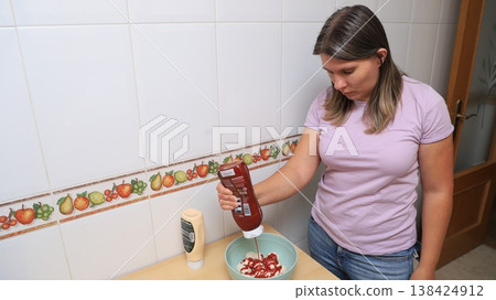 Woman in purple shirt pours ketchup onto a bowl of food in a kitchen Woman in purple shirt pours ketchup onto a bowl of food in a kitchen 138424912