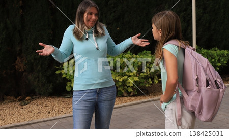 Loving mother and daughter face each other on paved path before school. Daughter gestures with hands while wearing pink backpack 138425031