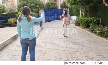 Mother waves goodbye to daughter at school gate, both smiling and ready for the day. Mother waves goodbye to daughter at school gate, both smiling and ready for the day. 138425053