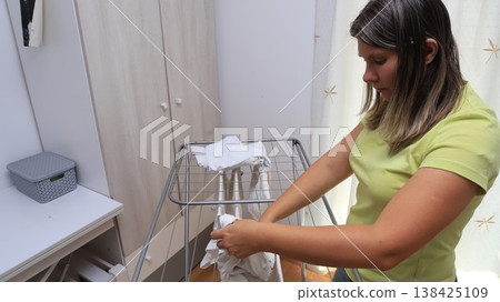 A woman is in a bright room, skillfully hanging laundry on a drying rack A woman is in a bright room, skillfully hanging laundry on a drying rack 138425109