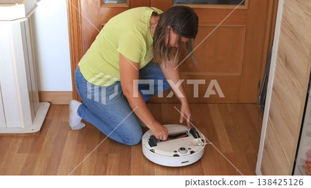 Woman repairs a robotic vacuum cleaner at home while kneeling on wooden floor near a door in daylight Woman repairs a robotic vacuum cleaner at home while kneeling on wooden floor near a door in daylight 138425126