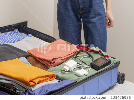 Travel gadgets and cables rest beside folded clothes during packing. Female hands place camera Travel gadgets and cables rest beside folded clothes during packing. Female hands place camera 138426926