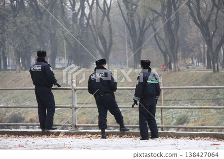 Three uniformed police officers stand by a riverside railing in a foggy park, observing the quiet surroundings. Three uniformed police officers stand by a riverside railing in a foggy park, observing the quiet surroundings. 138427064