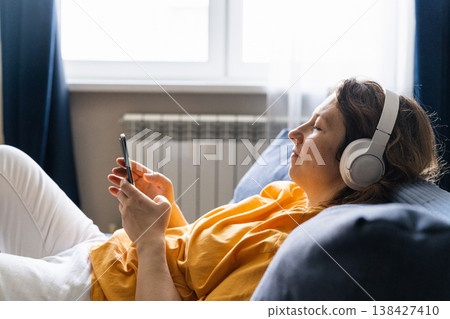 Woman lying on couch wearing white wireless headphones, enjoying audio from smartphone, surfing the internet in a cozy living room 138427410