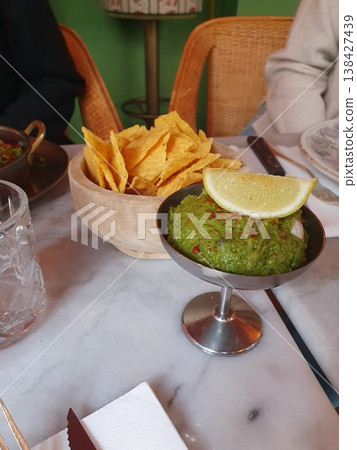 Fresh green guacamole topped with a lemon wedge served alongside crisp tortilla chips on a marble table in a cozy restaurant setting, evoking casual dining, sharing and social snacking. Fresh green guacamole topped with a lemon wedge served alongside crisp tortilla chips on a marble table in a cozy restaurant setting, evoking casual dining, sharing and social snacking. 138427439