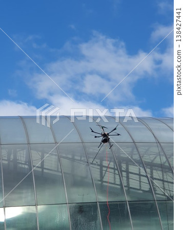 A tethered inspection drone hovers beside a curved glass roof of a modern building, conducting maintenance and window cleaning under a bright blue sky with scattered clouds, urban drone technology. A tethered inspection drone hovers beside a curved glass roof of a modern building, conducting maintenance and window cleaning under a bright blue sky with scattered clouds, urban drone technology. 138427441