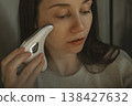 Young woman using microcurrent guasha face massager for lymphatic drainage and lifting, home beauty routine, skin care technology, pensive girl with electric massage tool, authentic self care. 138427632