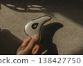 Hand holding modern microcurrent facial massager in warm sunlight with deep shadows, aesthetic beauty tech, minimalist skin care routine at home, sculptural guasha device, moody wellness concept. 138427750
