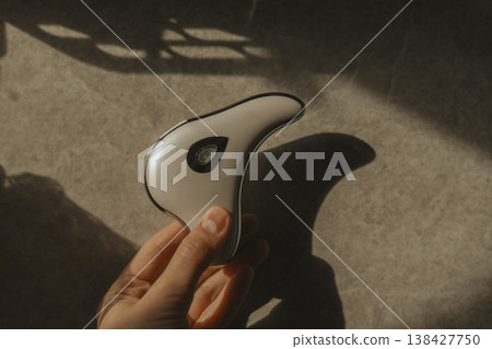 Hand holding modern microcurrent facial massager in warm sunlight with deep shadows, aesthetic beauty tech, minimalist skin care routine at home, sculptural guasha device, moody wellness concept. Hand holding modern microcurrent facial massager in warm sunlight with deep shadows, aesthetic beauty tech, minimalist skin care routine at home, sculptural guasha device, moody wellness concept. 138427750