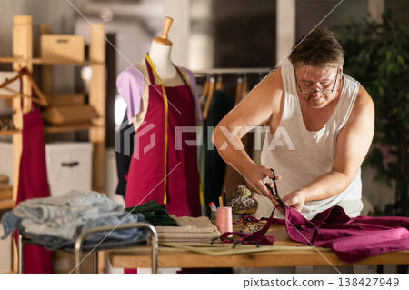 Middle-aged male dressmaker cutting textile in sewing workshop Middle-aged male dressmaker cutting textile in sewing workshop 138427949