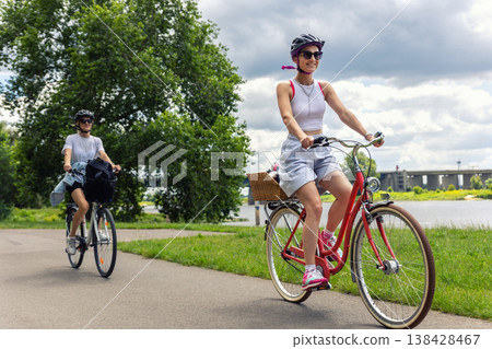 Women riding bicycles along riverside path during summer day with green park landscape and cloudy sky. Active lifestyle, friendship, outdoor recreation healthy activity travel freedom leisure cycling Women riding bicycles along riverside path during summer day with green park landscape and cloudy sky. Active lifestyle, friendship, outdoor recreation healthy activity travel freedom leisure cycling 138428467