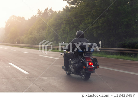 Motorcycle rider speeds along misty highway sunrise red tail light glow rain soaked asphalt drifting spray. Forest silhouette frames horizon truck fades fog evoking freedom adventure, risk solitude 138428497