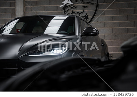 A matte black Lamborghini Urus sits in a low key lit workshop, LED headlight and hood reflecting softly, lion crest on cinderblock wall, blurred car foreground adds depth. 138429726