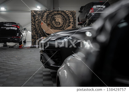 A matte black Lamborghini Urus stands in a dim workshop, with a Range Rover Autobiography and a Mercedes Benz Coupe AMG nearby, lifts, checker flooring, and a lion yin yang mural present. 138429757