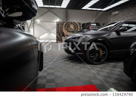 Matte black Lamborghini Urus stands right with bronze wheels under grid lights. Range Rover and Mercedes coupe sit behind near camo partition and crest on gray floor. Matte black Lamborghini Urus stands right with bronze wheels under grid lights. Range Rover and Mercedes coupe sit behind near camo partition and crest on gray floor. 138429790
