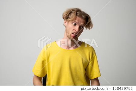 Young blonde man making a silly face and sticking out his tongue, expressing a humorous, playful, and childish emotion while looking to the side 138429795