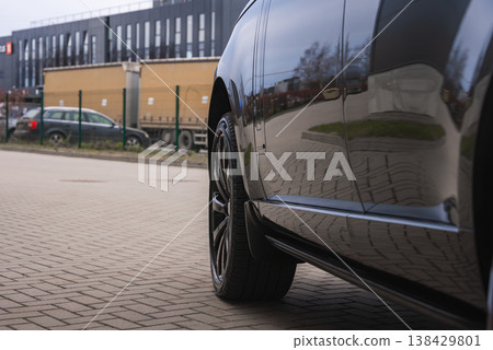 A glossy SUV side reflects trees, grass, a tan trailer truck, and a dark warehouse. A black alloy wheel fills the frame on a brick forecourt at a premium workshop. A glossy SUV side reflects trees, grass, a tan trailer truck, and a dark warehouse. A black alloy wheel fills the frame on a brick forecourt at a premium workshop. 138429801