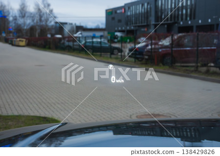 A luxury car HUD projects 0 kmh on a head up display and a speed limit icon on the windshield while idling by service bays and parked cars at a modern complex. A luxury car HUD projects 0 kmh on a head up display and a speed limit icon on the windshield while idling by service bays and parked cars at a modern complex. 138429826