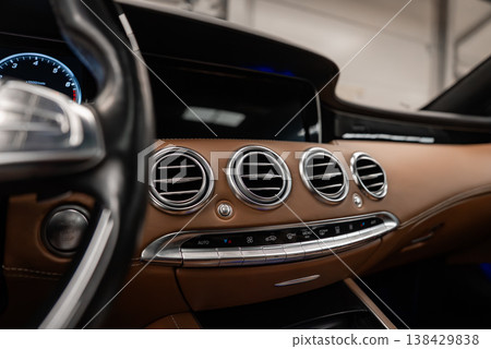 Close up of a tan leather dashboard with four chrome vents, analog clock, wide black infotainment screen, and slim metallic controls indoors under soft diffused light. Close up of a tan leather dashboard with four chrome vents, analog clock, wide black infotainment screen, and slim metallic controls indoors under soft diffused light. 138429838