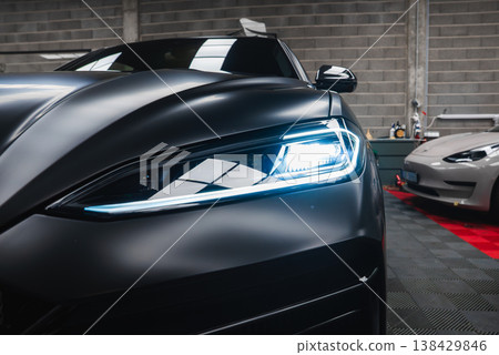 Close up of left Lamborghini Urus SE LED headlight in a premium workshop. A silver Tesla Model 3 sits on a red mat with detailing gear and checkered floor. Close up of left Lamborghini Urus SE LED headlight in a premium workshop. A silver Tesla Model 3 sits on a red mat with detailing gear and checkered floor. 138429846