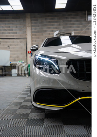 Close view shows Panamericana grille, LED headlight, and yellow bumper line on a Mercedes AMG in an auto workshop with brick walls, fluorescent panels, and checkered floor. Close view shows Panamericana grille, LED headlight, and yellow bumper line on a Mercedes AMG in an auto workshop with brick walls, fluorescent panels, and checkered floor. 138429851