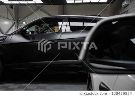 A matte black Lamborghini Urus stands in a modern workshop, checkered floor beneath, with a white AMG coupe foreground detail, grid lights above, subdued indoor lighting. 138429854