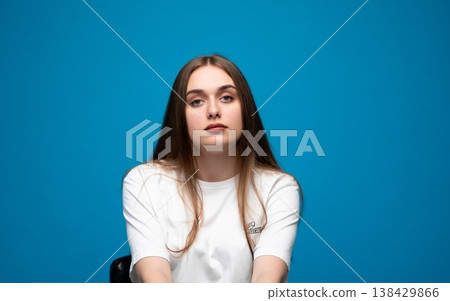Young woman with long brown hair posing confidently in a white t-shirt against a vibrant blue studio background 138429866