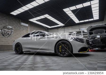 A white Mercedes AMG coupe sits on black wheels with yellow calipers in a high end workshop, checker flooring and bright panels, with another car hood open nearby. A white Mercedes AMG coupe sits on black wheels with yellow calipers in a high end workshop, checker flooring and bright panels, with another car hood open nearby. 138429954