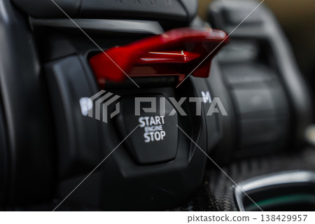 Close up macro shows red flip up cover revealing START ENGINE STOP button on a luxury Lamborghini Urus SE console, with rotary controls and carbon fiber trim under moody indoor light. Close up macro shows red flip up cover revealing START ENGINE STOP button on a luxury Lamborghini Urus SE console, with rotary controls and carbon fiber trim under moody indoor light. 138429957
